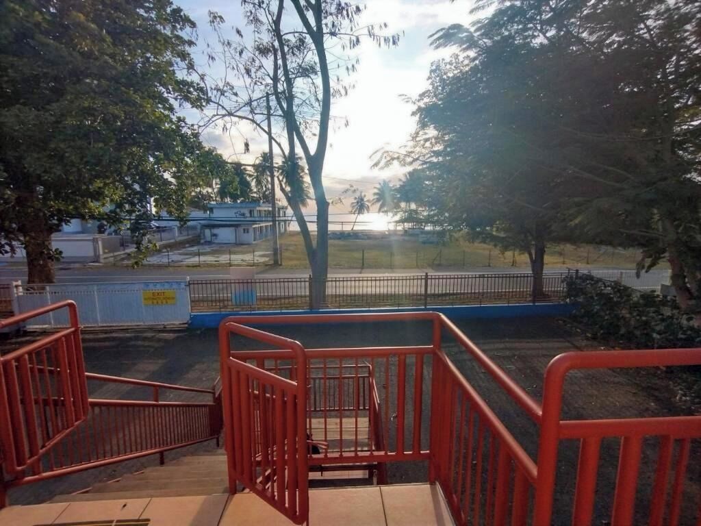 Un espacio exterior cercado con una escalera que baja, árboles y un edificio al fondo.