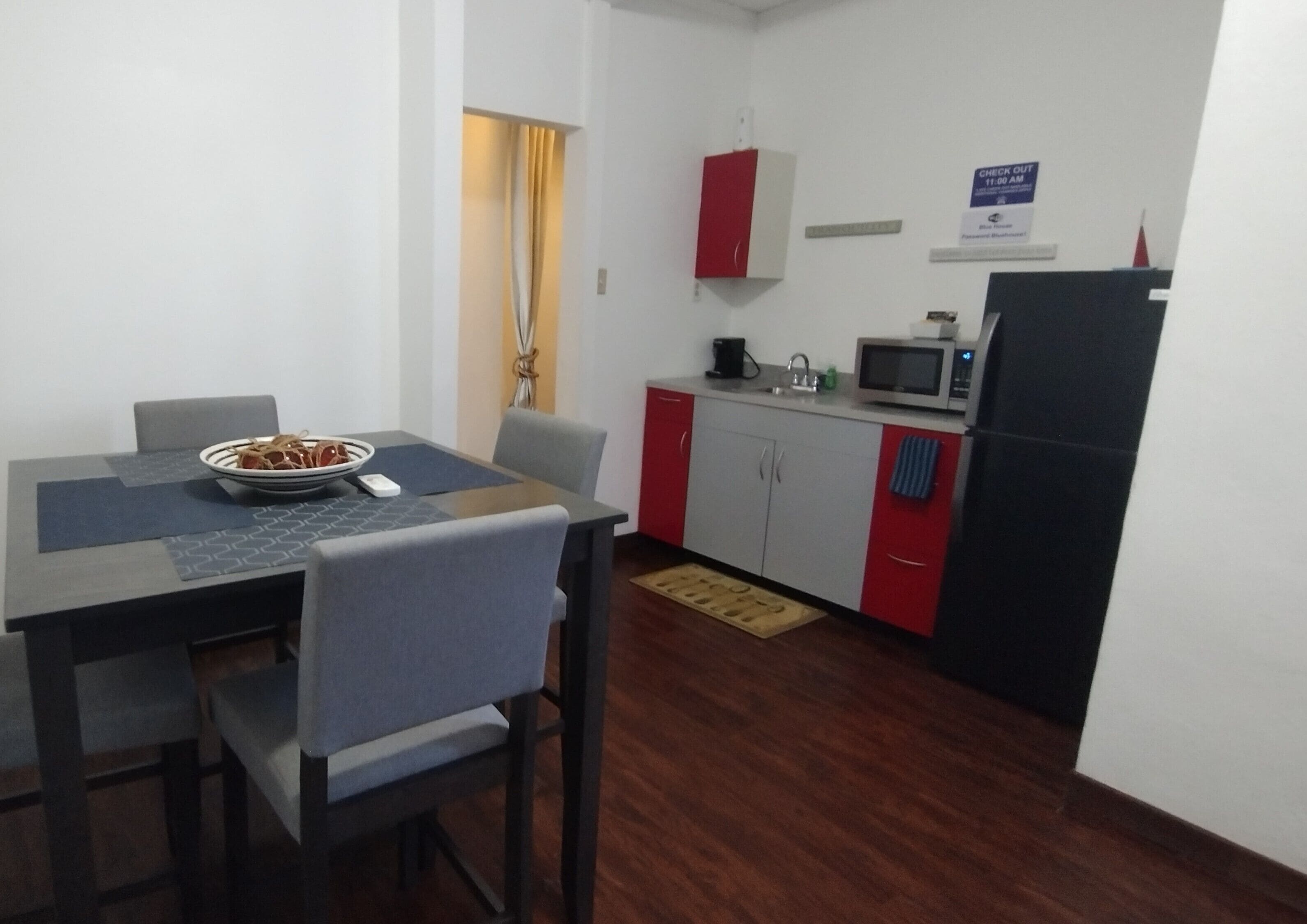 Cocina compacta con microondas, pileta y muebles color rojo. Hay una mesa de comedor con sillas y un cuenco de frutas.