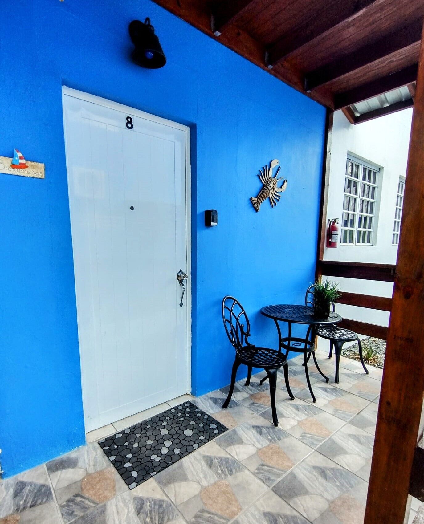Un mur extérieur bleu avec une porte blanche, une petite table d’extérieur avec deux chaises et un élément décoratif mural.