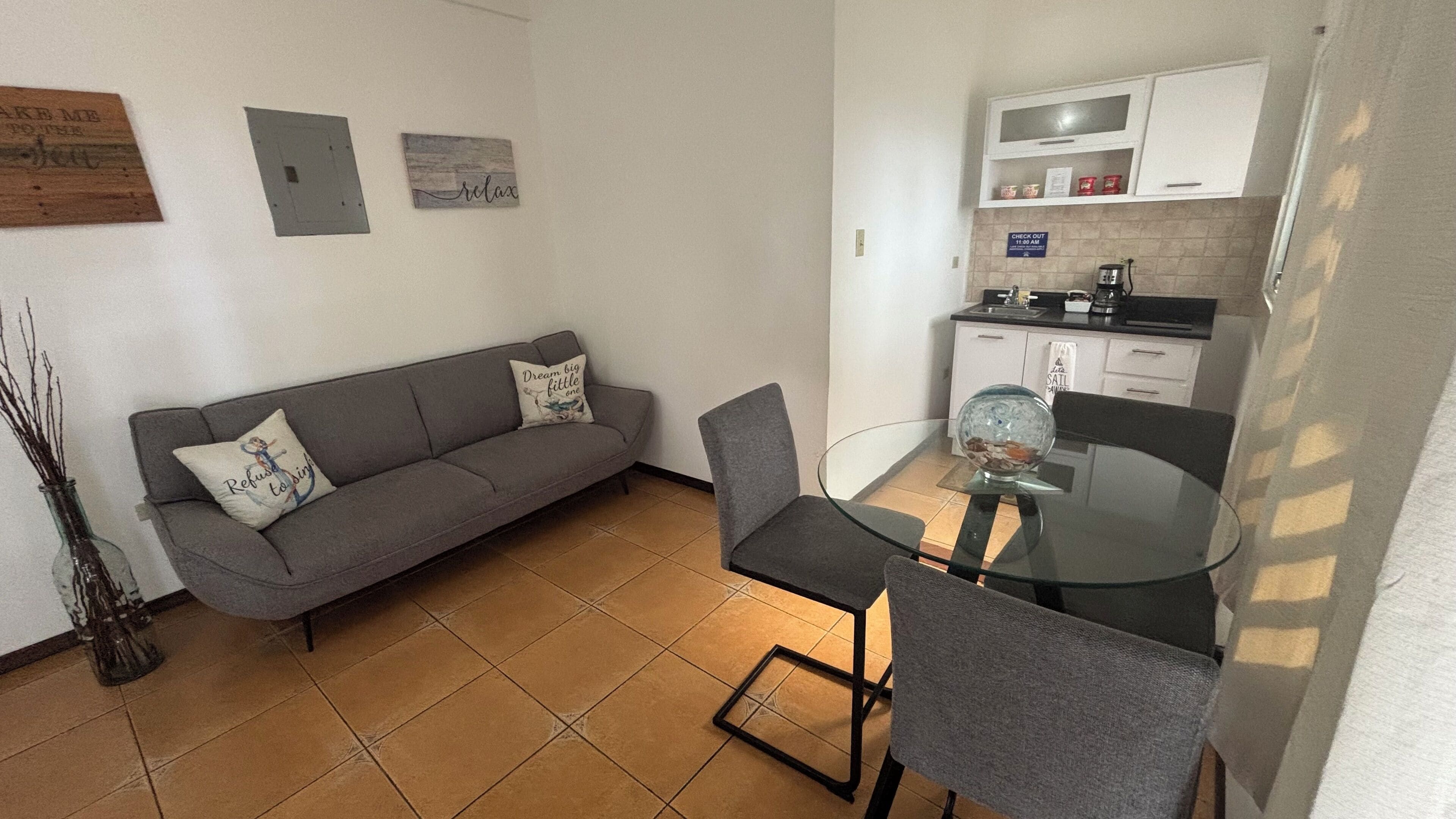 Ein kompakter Wohnbereich mit grauem Sofa, einem Glastisch und einer Küchenzeile mit weißen Schränken und einer Kaffeemaschine.