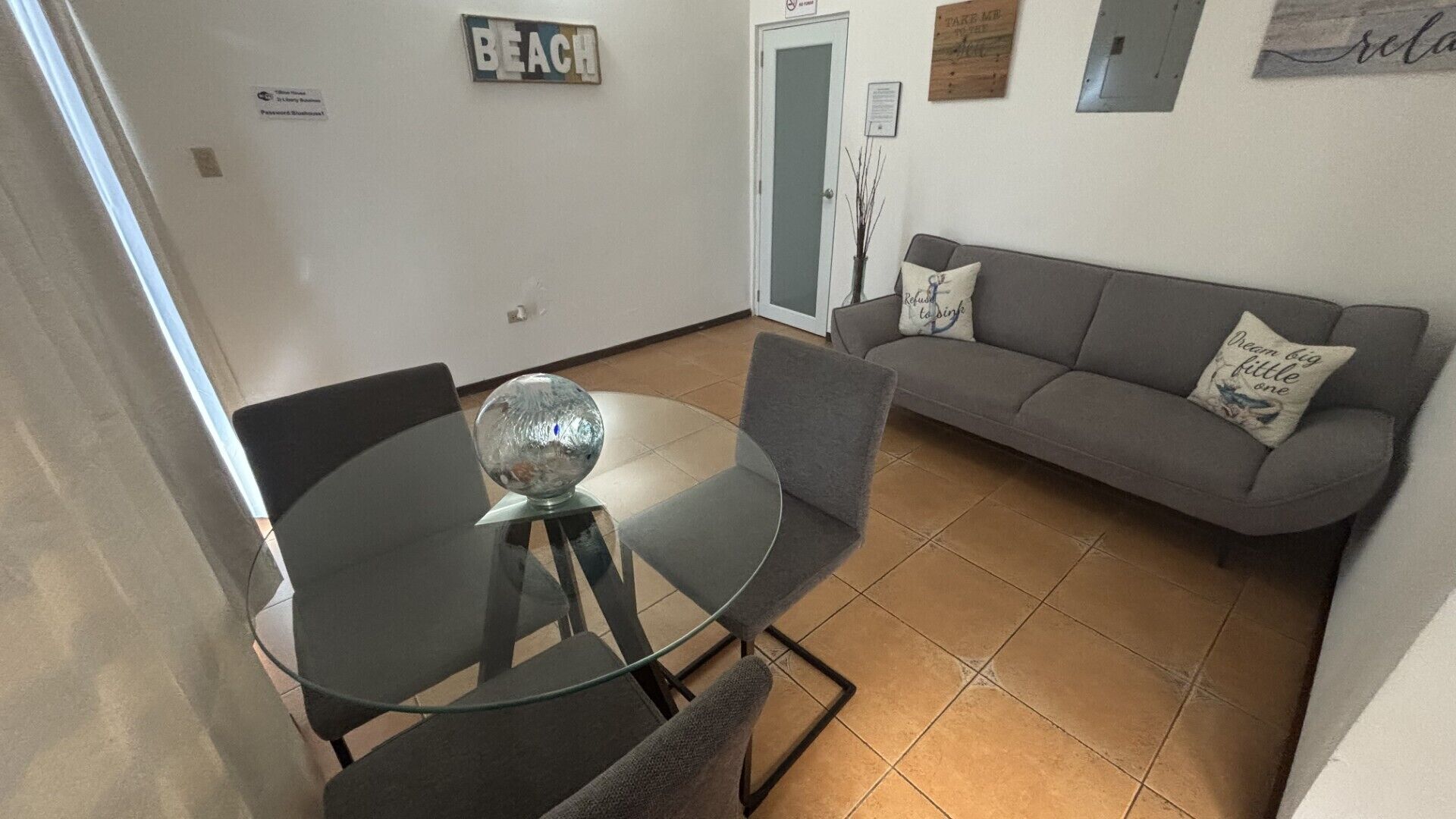 Un'area pranzo con tavolo e sedie in vetro, un divano grigio con cuscini e un cartello a parete con su scritto 'BEACH'.
