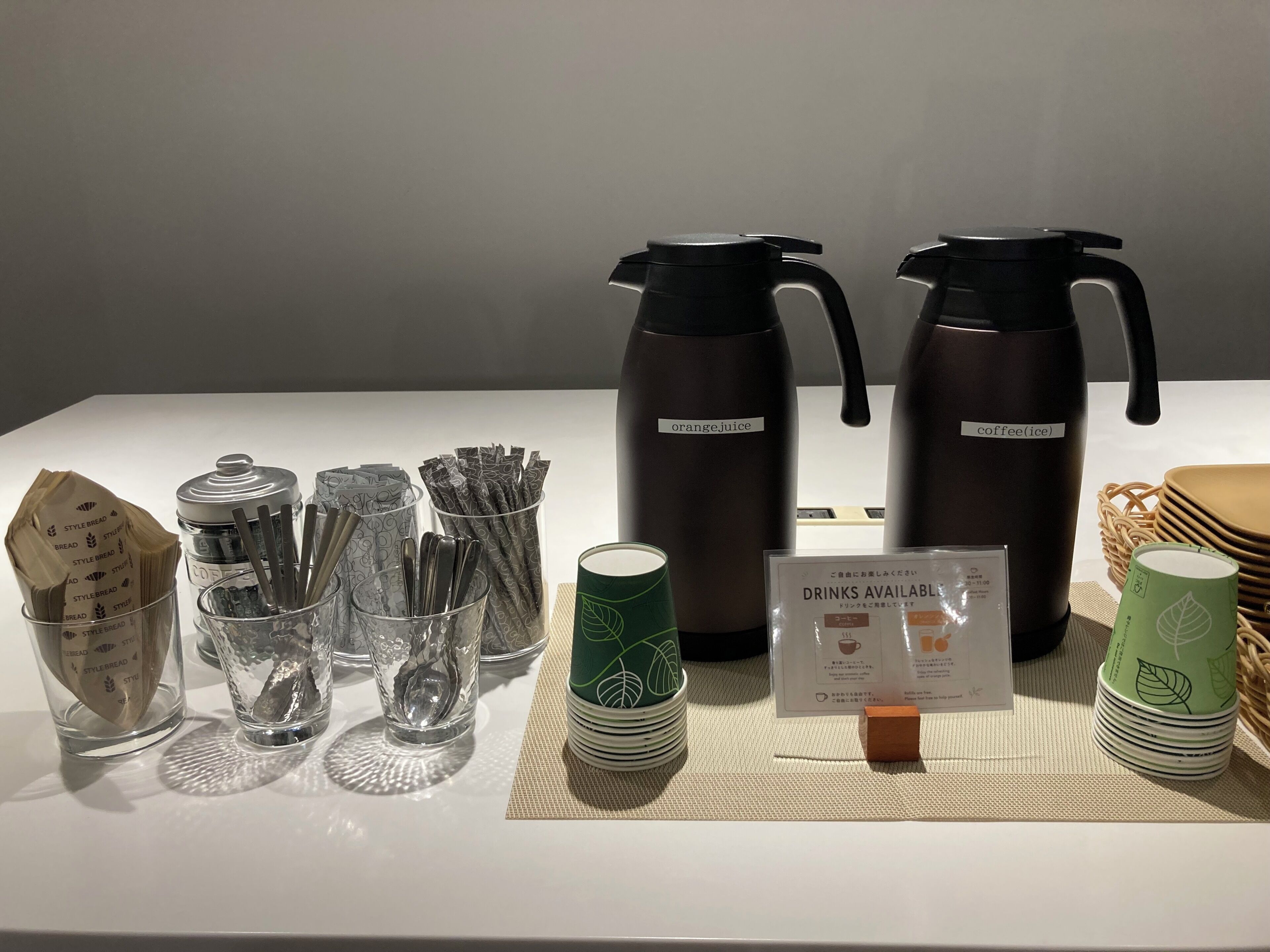 Un point de distribution de boissons comprenant deux thermos, un panneau indiquant les boissons disponibles et une pile de gobelets jetables.