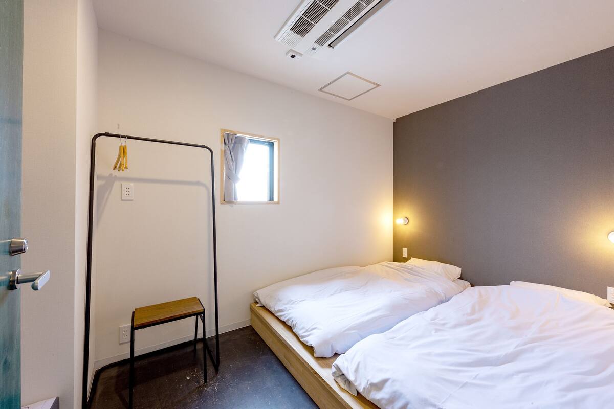 Une petite chambre avec un lit simple, un banc en bois, une fenêtre avec un rideau et un luminaire fixé au mur.