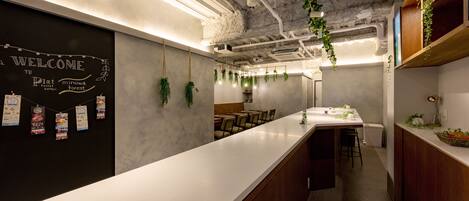 A modern kitchen with a long counter, hanging plants, and a chalkboard with a welcome message.
