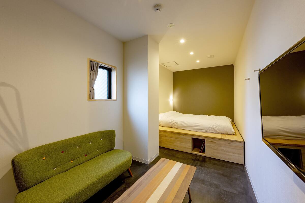 Une petite pièce moderne avec un canapé vert, un lit doté d’une tête de lit en bois et un miroir au mur.
