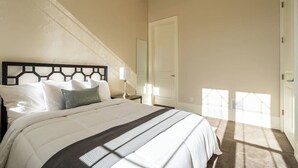 Quarto com uma cama grande, mesa de cabeceira com abajur, porta e janela com luz do sol.