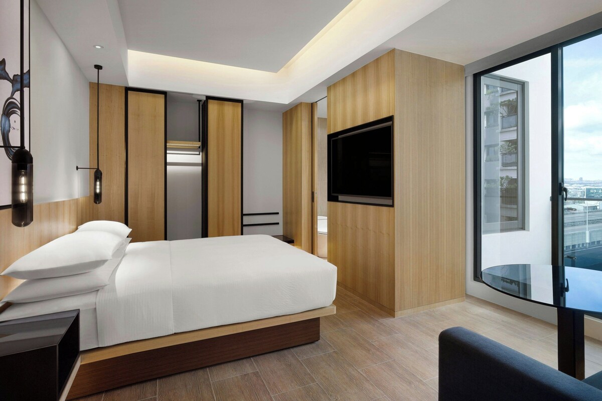 A modern hotel room with a bed, a TV, and a large window offering a city view.