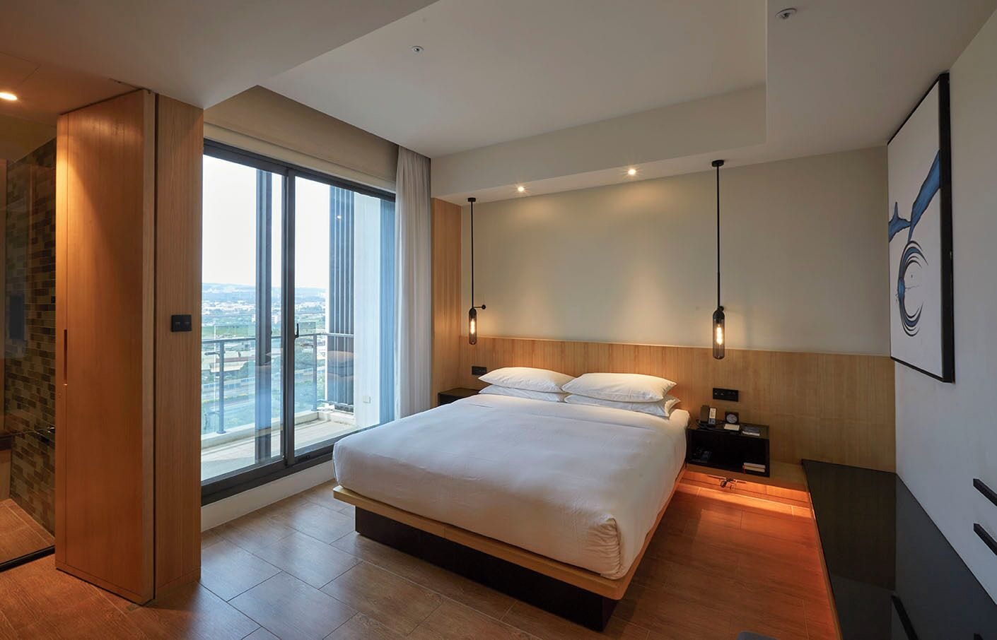 Habitación de hotel moderna con una cama grande, una mesita de noche, un escritorio y un amplio ventanal con vista a la ciudad.