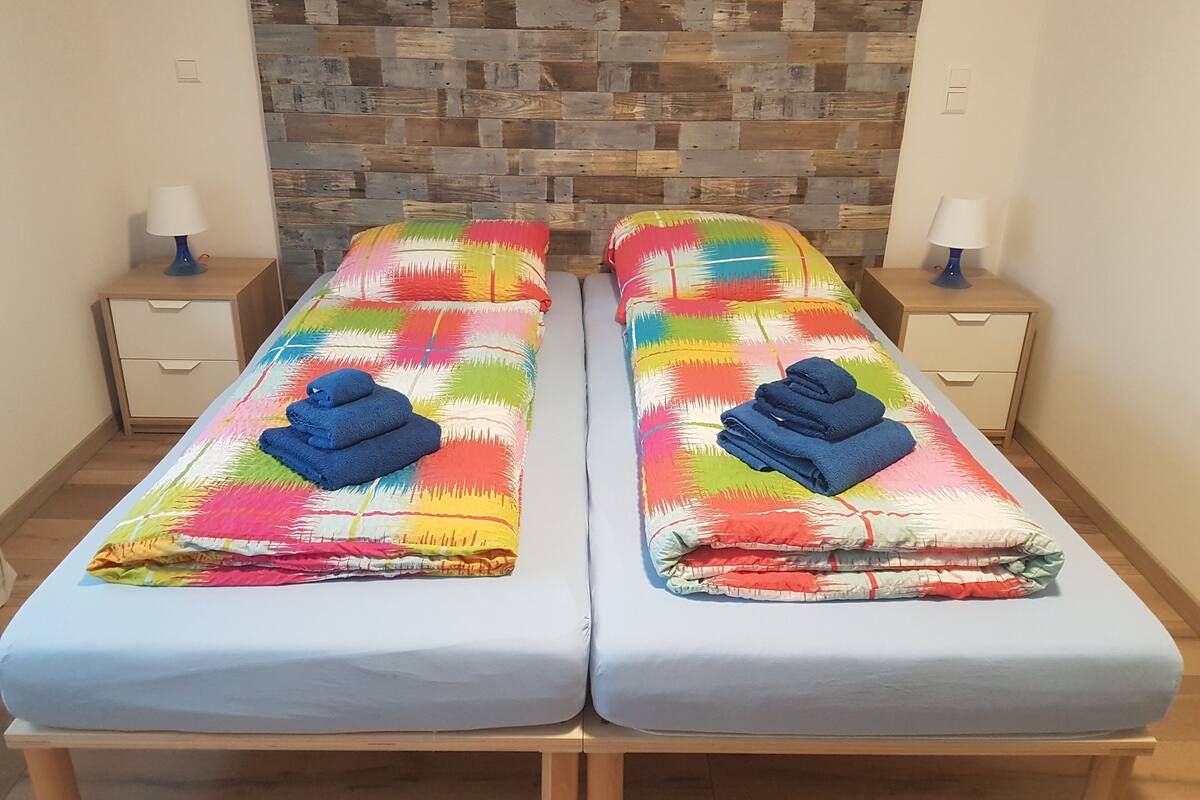 Dos camas individuales con ropa de cama y toallas coloridas, mesitas de noche de madera y un cabecero con estampado de piedra.