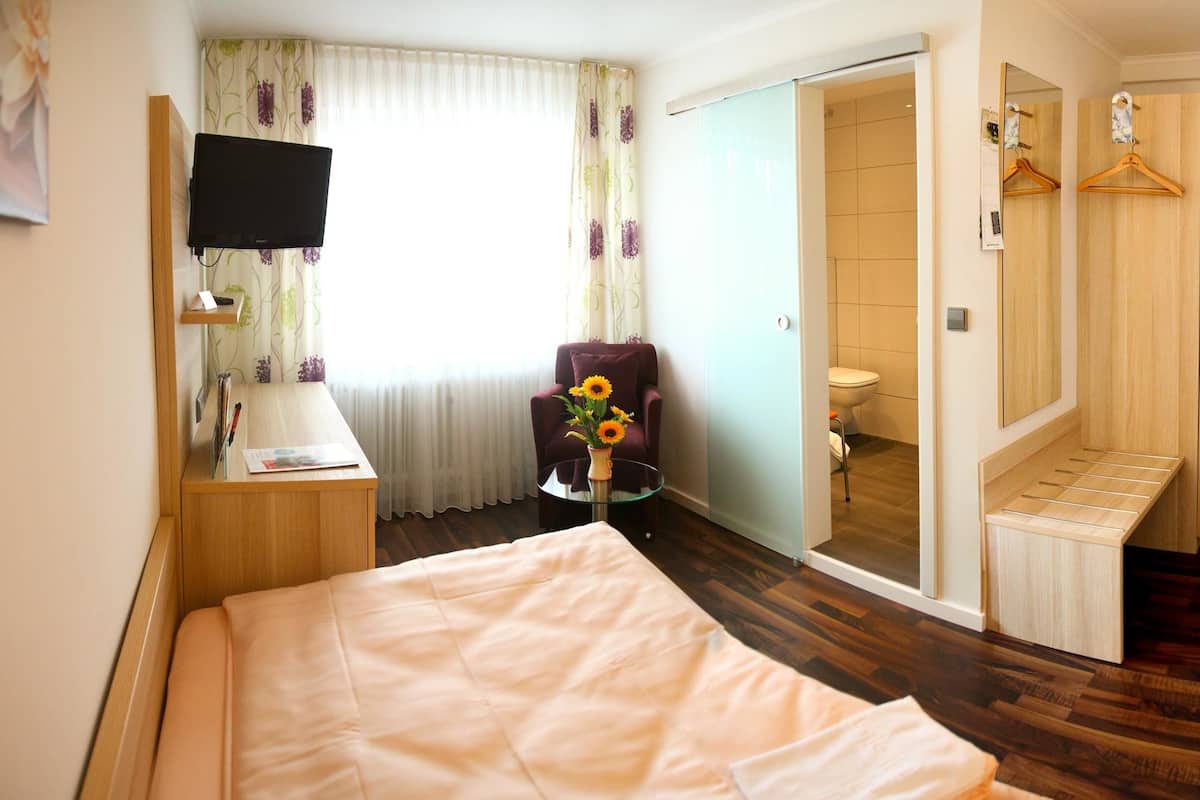Ein Hotelzimmer mit einem Bett, einem Schreibtisch mit Lampe, einem Sessel mit Blumenvase, einem Fernseher an der Wand, einem Badezimmer mit Toilette und einem Spiegel mit Handtuch.
