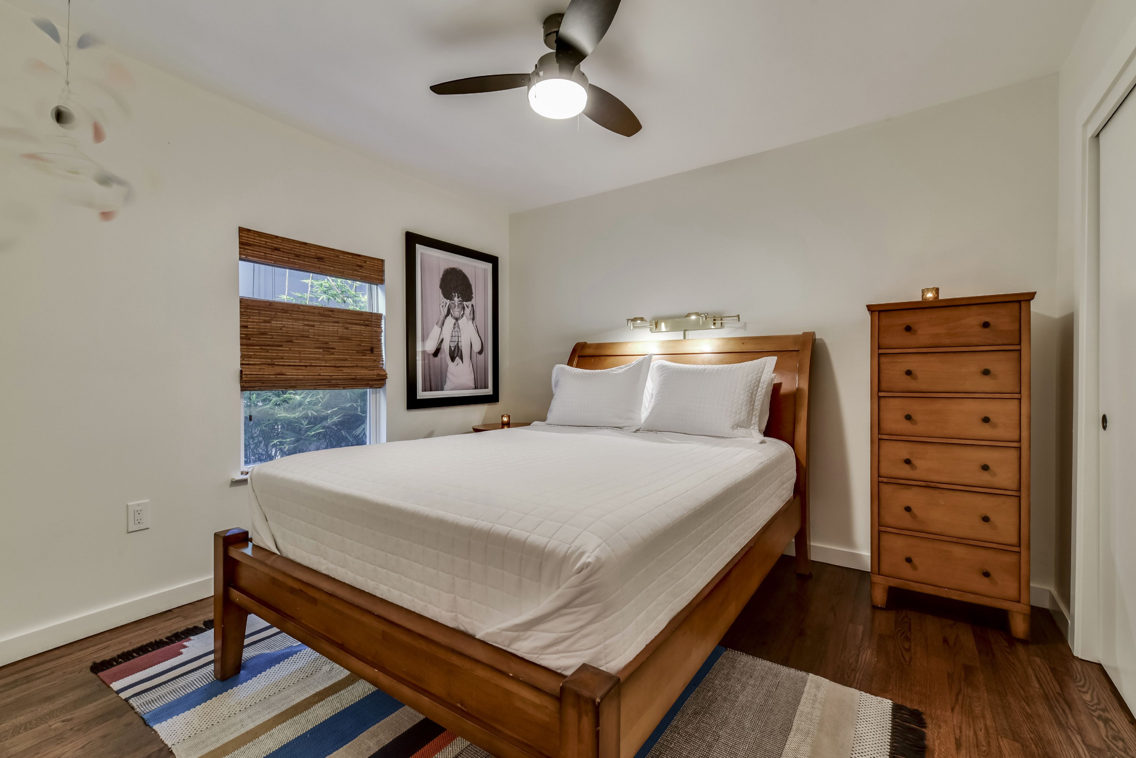 A bedroom with a wooden bed, a ceiling fan, a nightstand, a dresser, and a window with blinds.