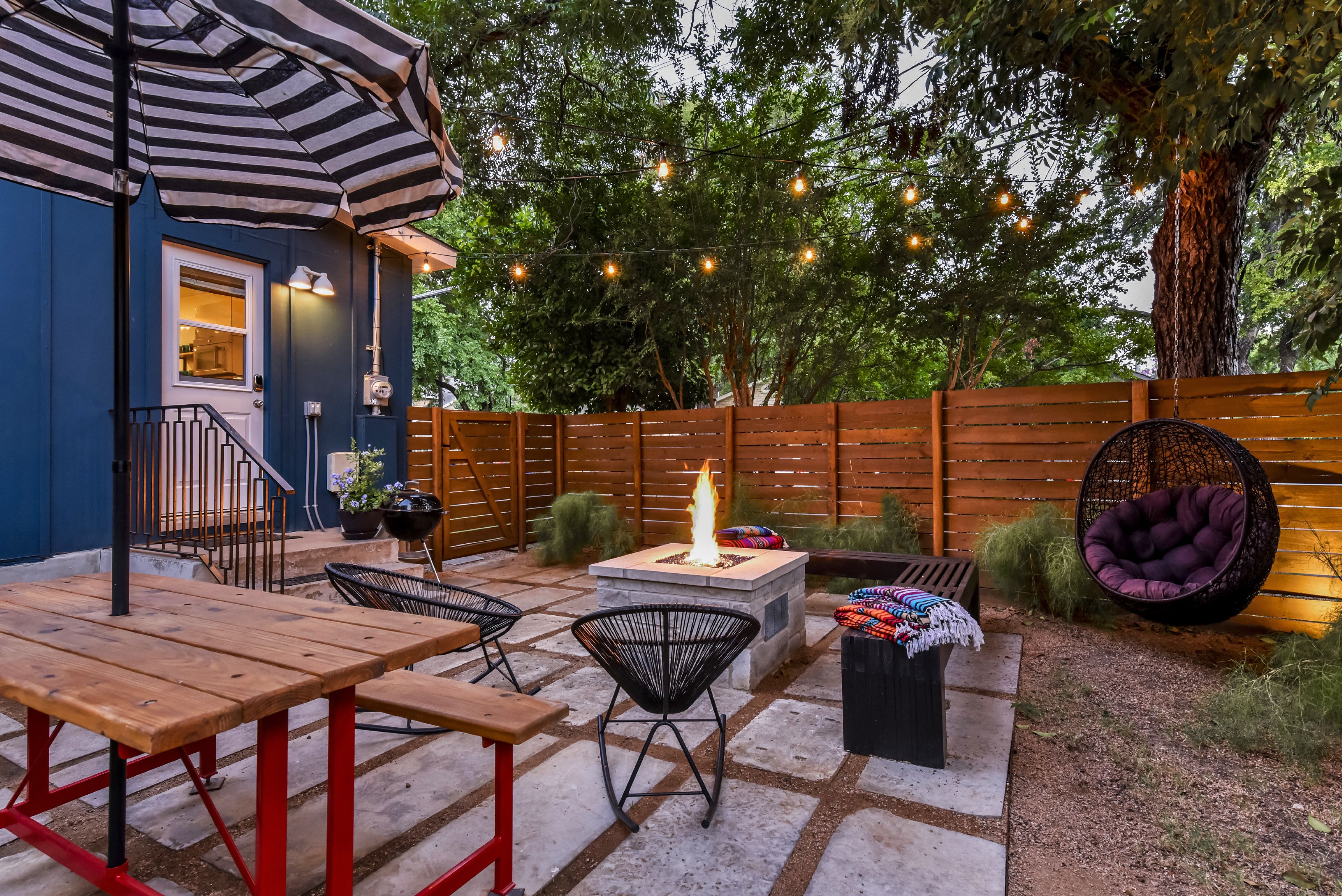 An outdoor patio with a wooden table, chairs, a fire pit, and string lights.