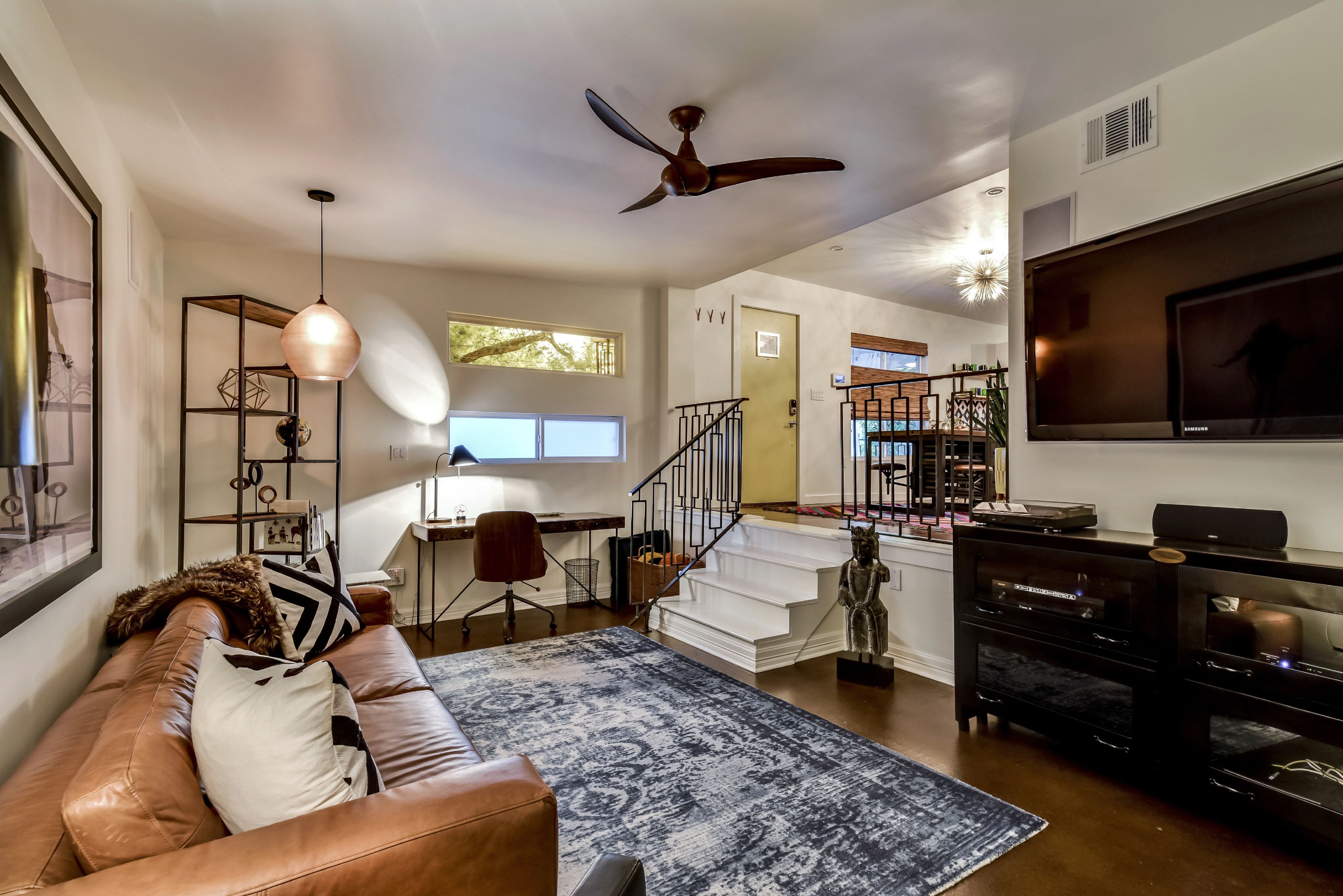 A modern living room with a leather sofa, a patterned rug, a ceiling fan, a TV, and a staircase leading to an upper level.