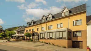 A yellow building with the name "Zur Neeroburg" on the side, featuring a brick facade, multiple windows, and a covered outdoor seating area. - Zur Neroburg (Neroth)