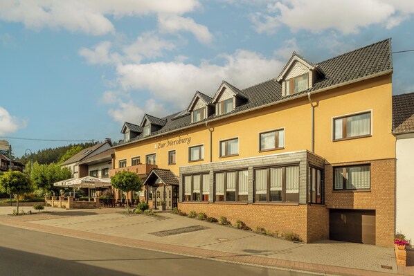 A yellow building with the name "Zur Neeroburg" on the side, featuring a brick facade, multiple windows, and a covered outdoor seating area. - Zur Neroburg (Neroth)