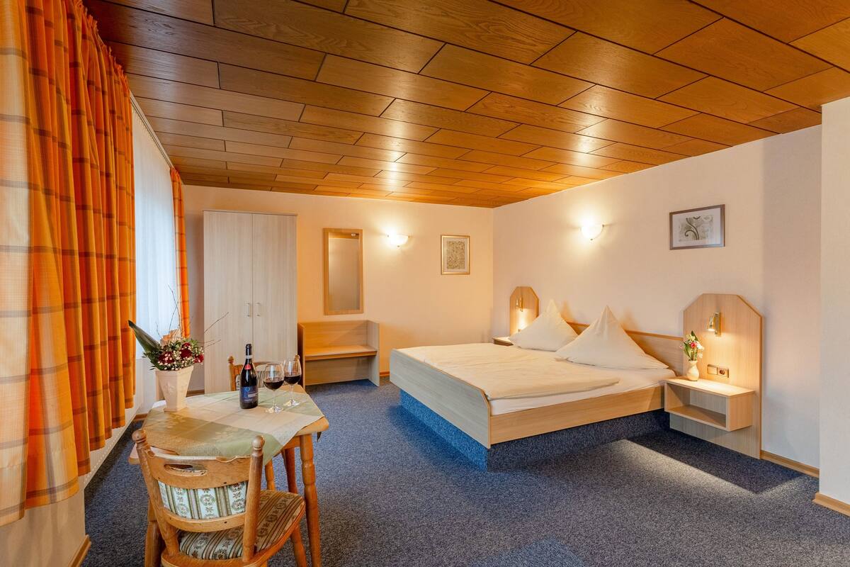 Habitación de hotel con techo de madera, cama, escritorio con silla, pequeña mesa con un florero y vino, y mesita de noche con lámpara.