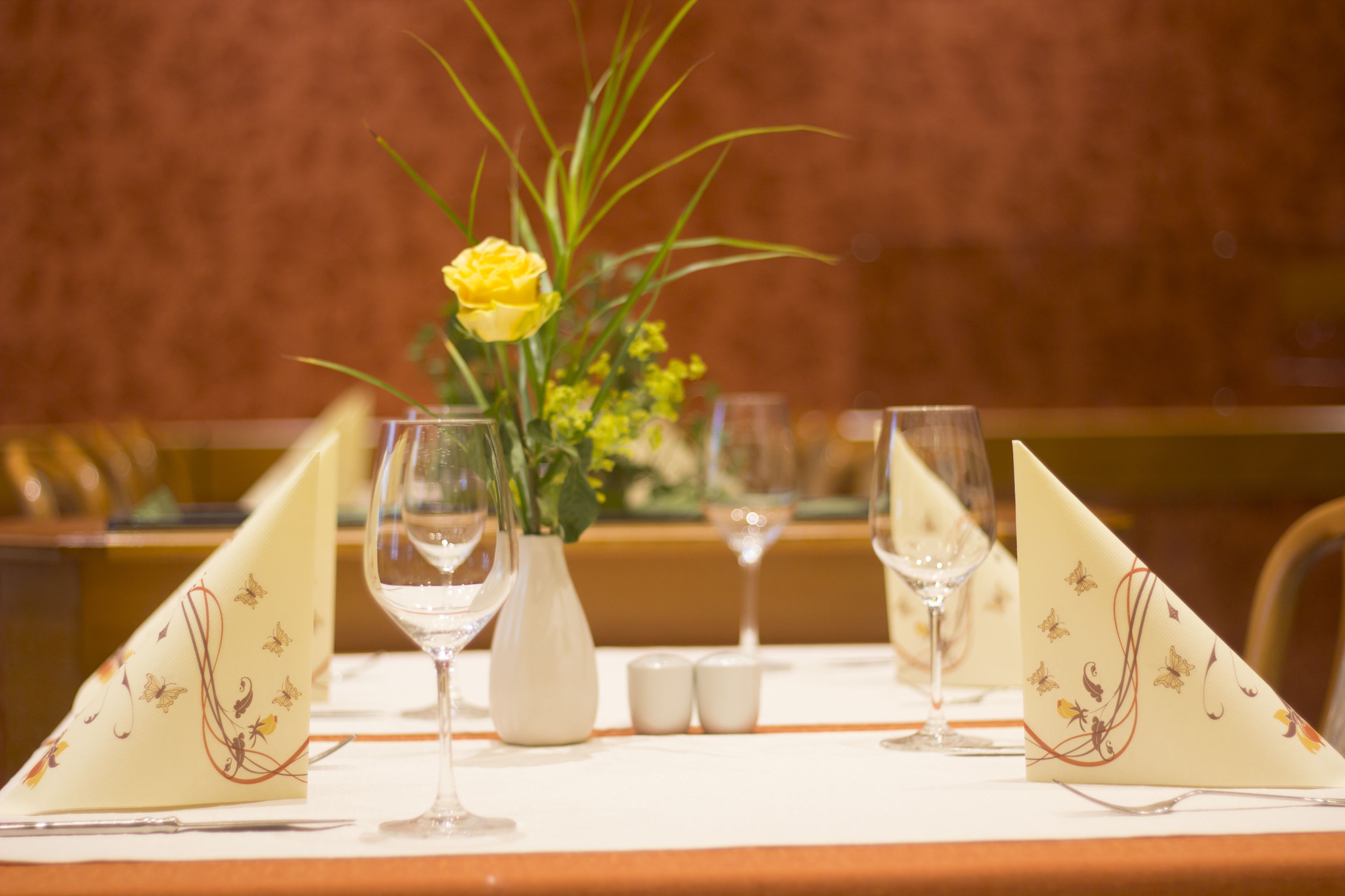 Una mesa de comedor dispuesta con servilletas blancas, un florero con flores amarillas y dos copas de vino.