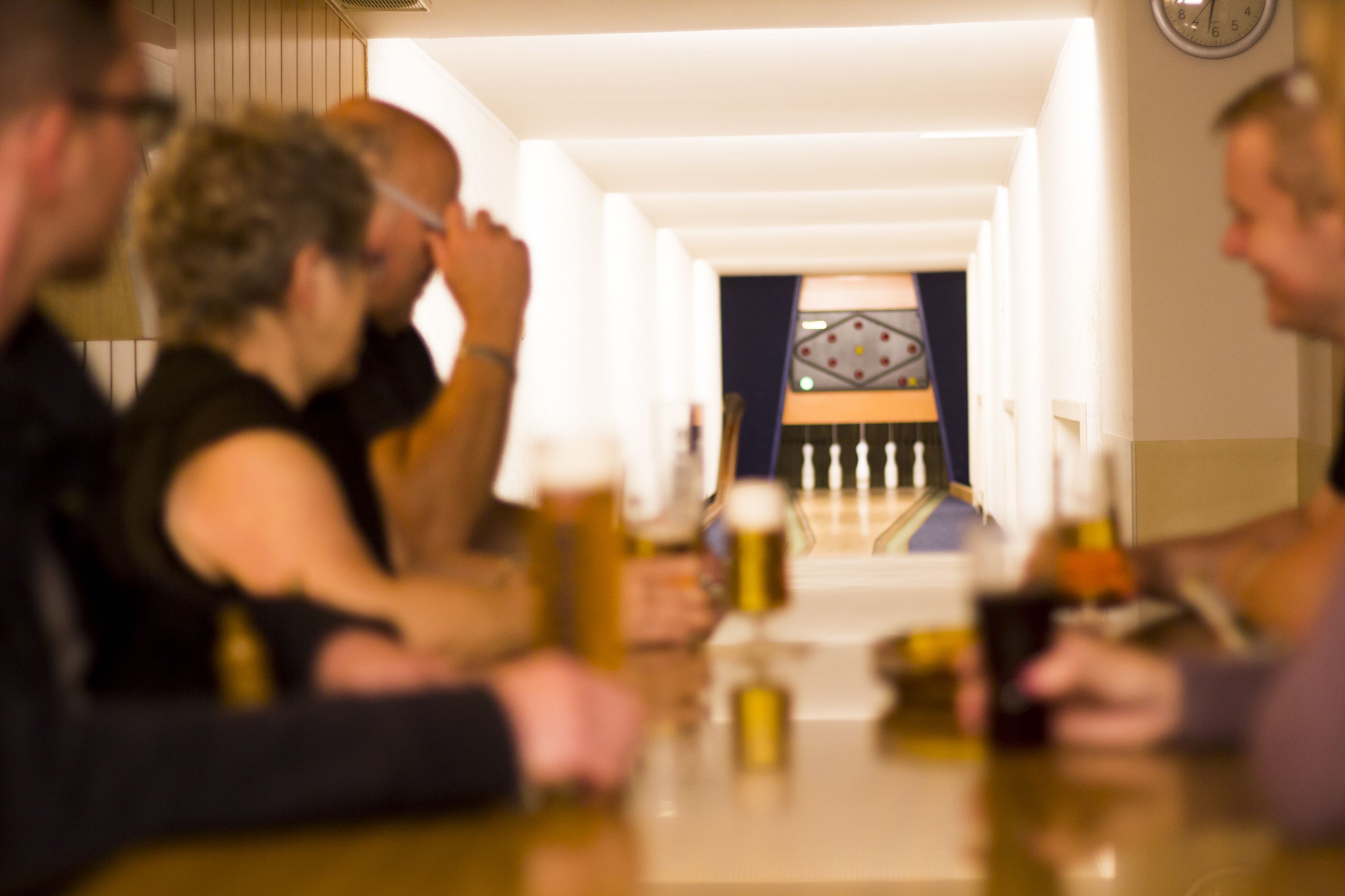 A group of people seated at a table with drinks, in a room with a clock on the wall and a patterned curtain in the background.