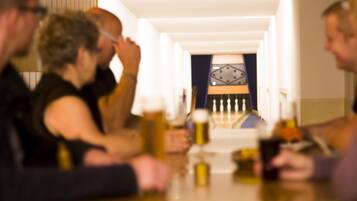 A group of people seated at a table with drinks, in a room with a clock on the wall and a patterned curtain in the background.
