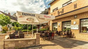 An outdoor seating area with tables and chairs, a large umbrella with branding, and a brick building in the background. - Zur Neroburg (Neroth)