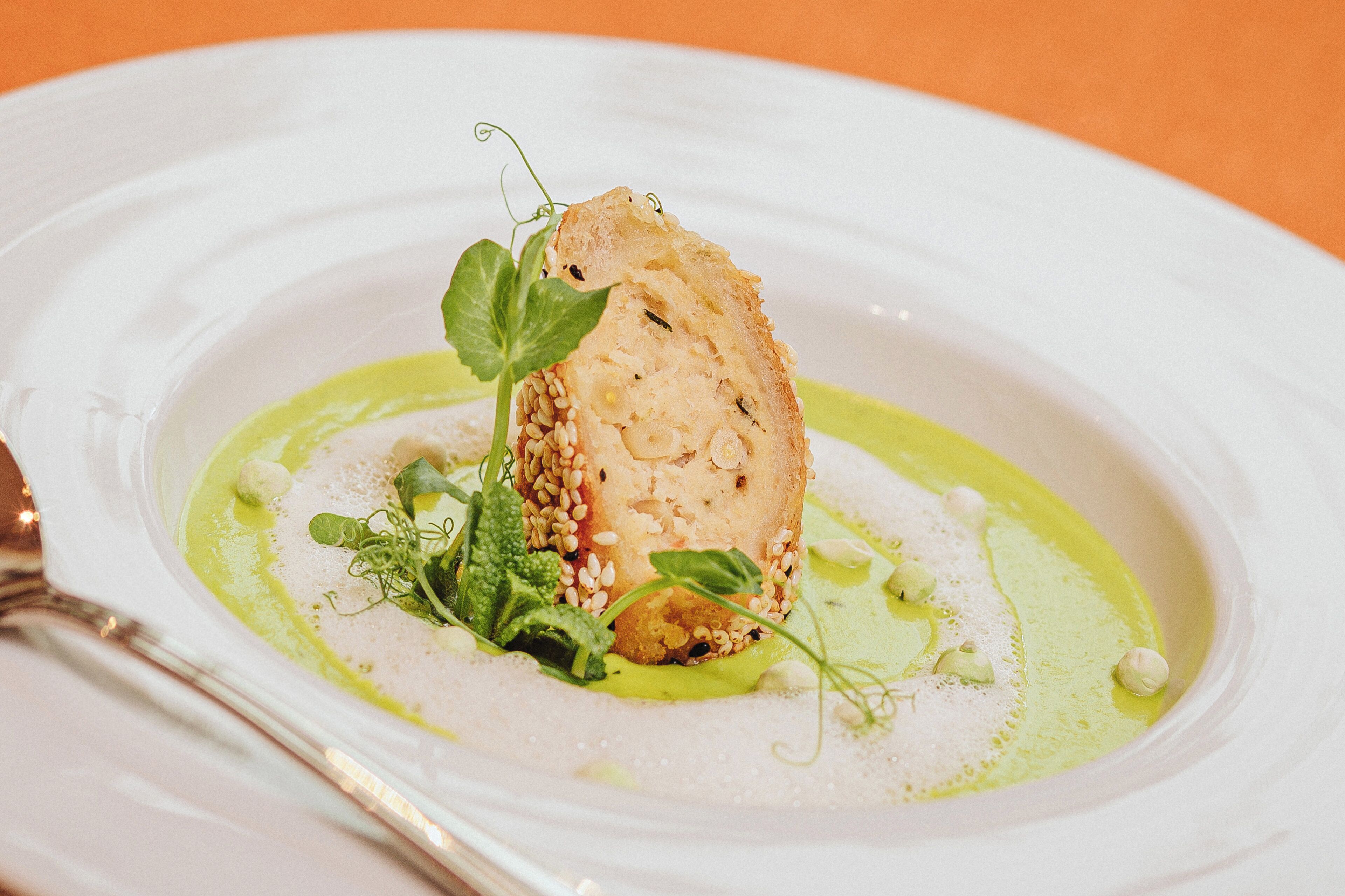 A white plate with a piece of food garnished with green leaves and sesame seeds, accompanied by a green sauce.