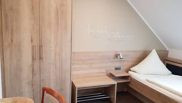 A bedroom with a wooden wardrobe, a desk, a bed, and a wall-mounted light.