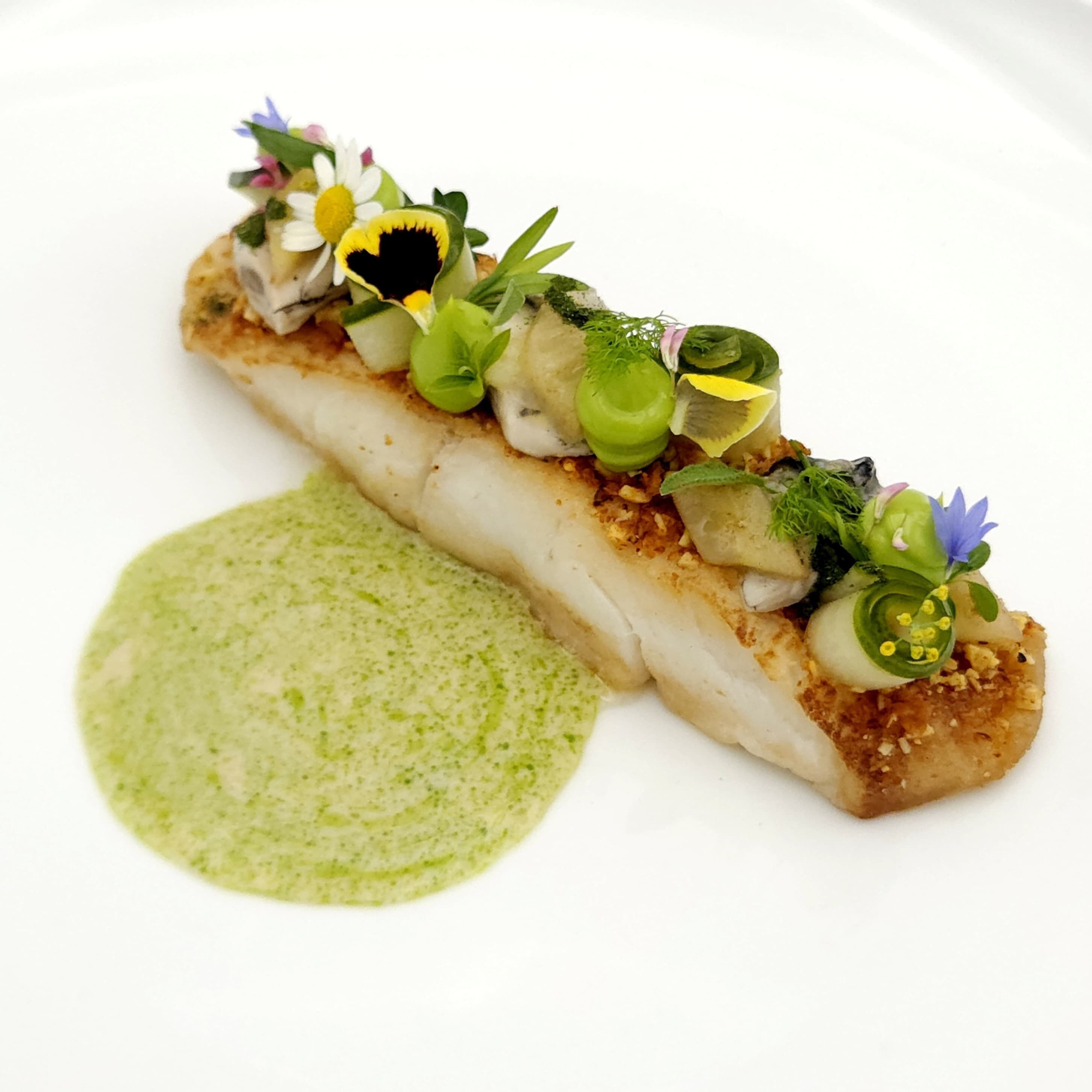 Un plato con salsa verde y un trozo de pescado cubierto de verduras y flores.