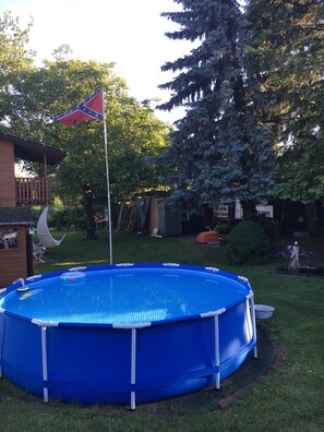Une piscine bleue hors sol, surmontée d’un mât pour drapeau, entourée d’une terrasse en bois et d’une pelouse.