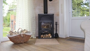 A wood-burning stove with a basket of firewood in front, a white chair, and a view of the outdoors through a window. - Haus Elbromantik (Sandau)