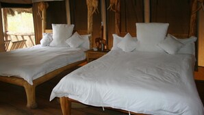 Two wooden beds with white linens, a wooden nightstand with a lamp, and a thatched roof structure. - Roika Tarangire Tented Lodge (Tarangire National Park)