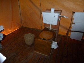 Una habitación pequeña con suelo de madera, un inodoro sencillo, una cesta tejida pequeña y un refrigerador blanco.