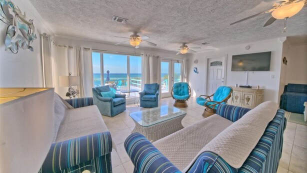 Amplia sala de estar con un sofá grande, dos sillones, una mesa de centro y una televisión. La habitación tiene ventanas amplias con vistas a la playa.