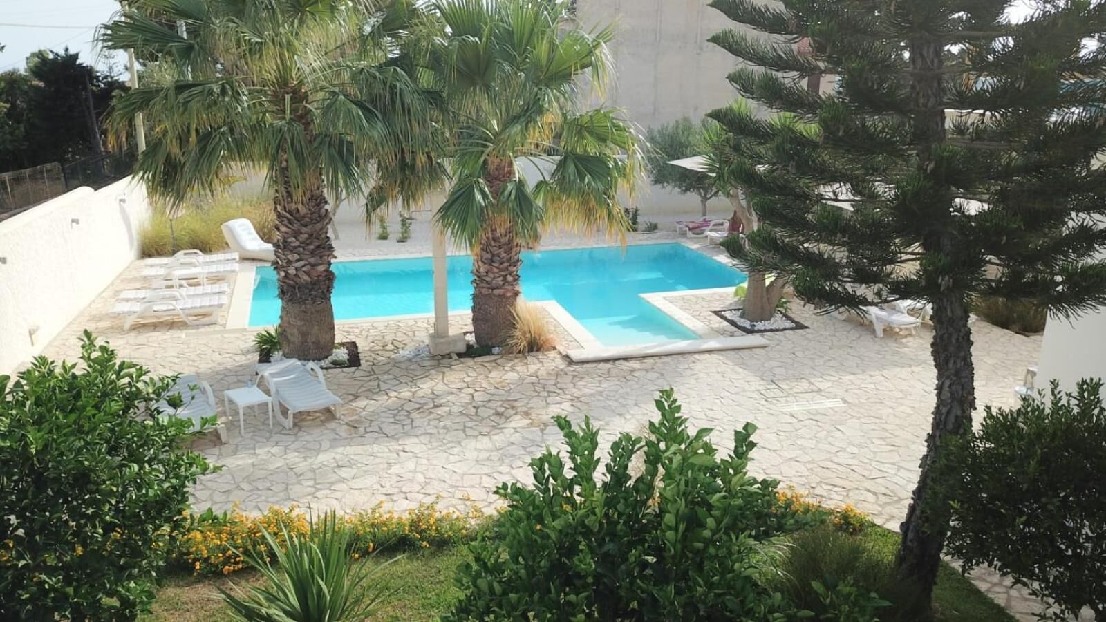 Área de piscina com espreguiçadeiras, palmeiras e um pátio com piso de pedra.