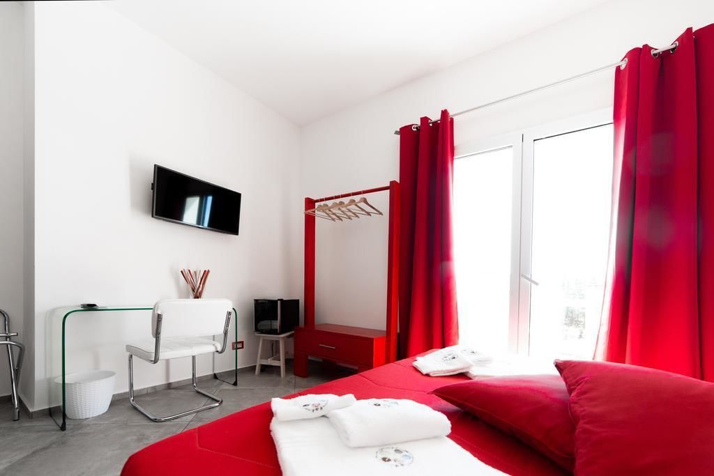 A hotel room with a bed, a TV mounted on the wall, a desk with a chair, a small table, and a red curtain.