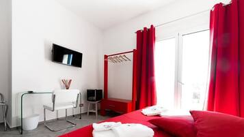 A hotel room with a bed, a TV mounted on the wall, a desk with a chair, a small table, and a red curtain.