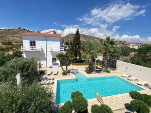 Un edificio de dos pisos con piscina, rodeado de palmeras y un paisaje montañoso.