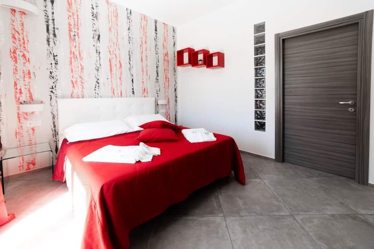 A bedroom with a bed, red bedding, a white headboard, a red wall decoration, a door, and a wall-mounted shelf.