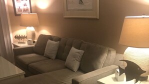 Ett vardagsrum med en hörnsoffa, ett litet bord med en lampa och en inramad tavla på väggen.