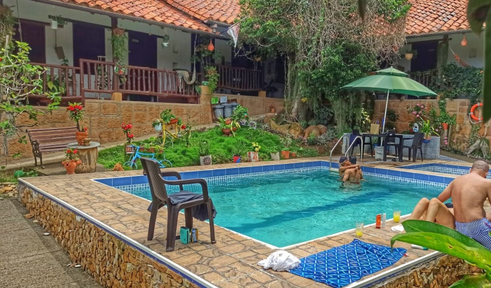 Una piscina con borde de azulejos, rodeada de muros de piedra y plantas en macetas. Hay zonas de estar al aire libre y un porche cubierto con sombrillas.