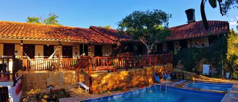Zona de piscina con muro de piedra y terraza de madera, rodeada de edificios con techos de tejas rojas.