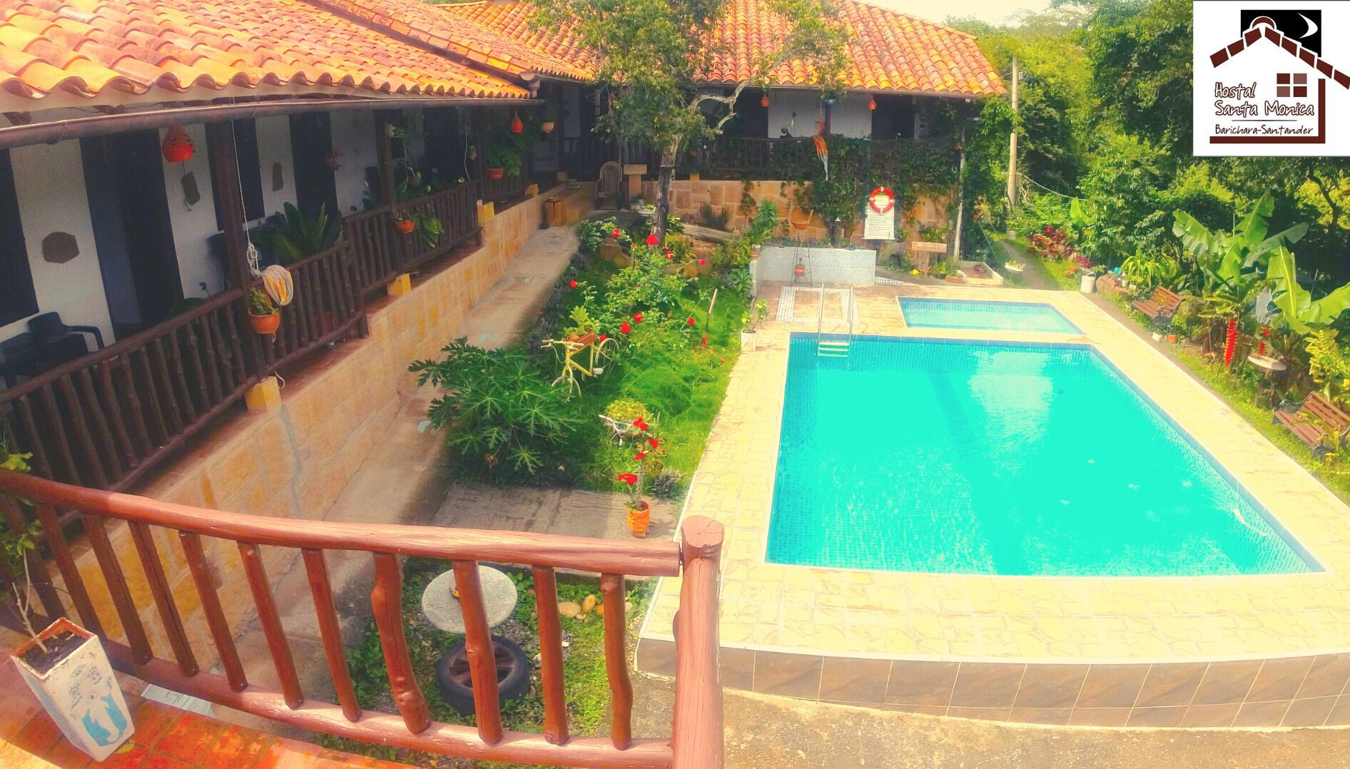 Uma área de piscina com um deck ladrilhado, uma cerca de madeira e uma placa para Hostal Santa Monica ao fundo.