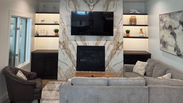 A modern living room with a fireplace, a large flat-screen TV mounted on a marble wall, a grey sofa, a brown leather armchair, and a small side table with decorative items. - Lakefront resort, pool, tennis, pickleball with a private beach, fast EV charger (Whitefish)