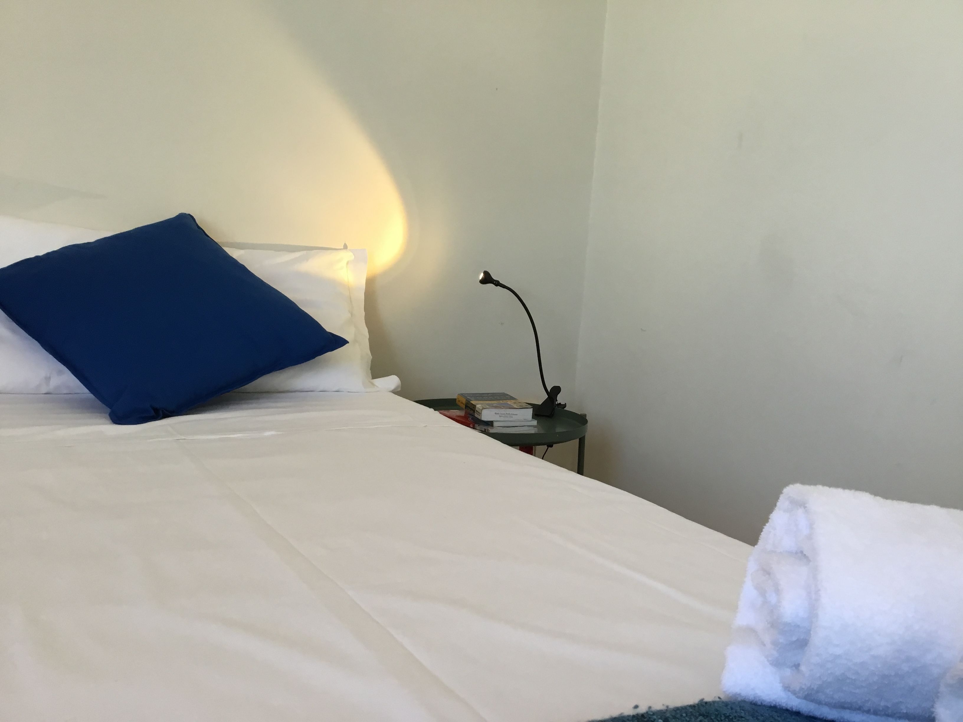 A neatly made bed with white linens and a blue pillow, a bedside lamp, and a small table with a book and a phone.