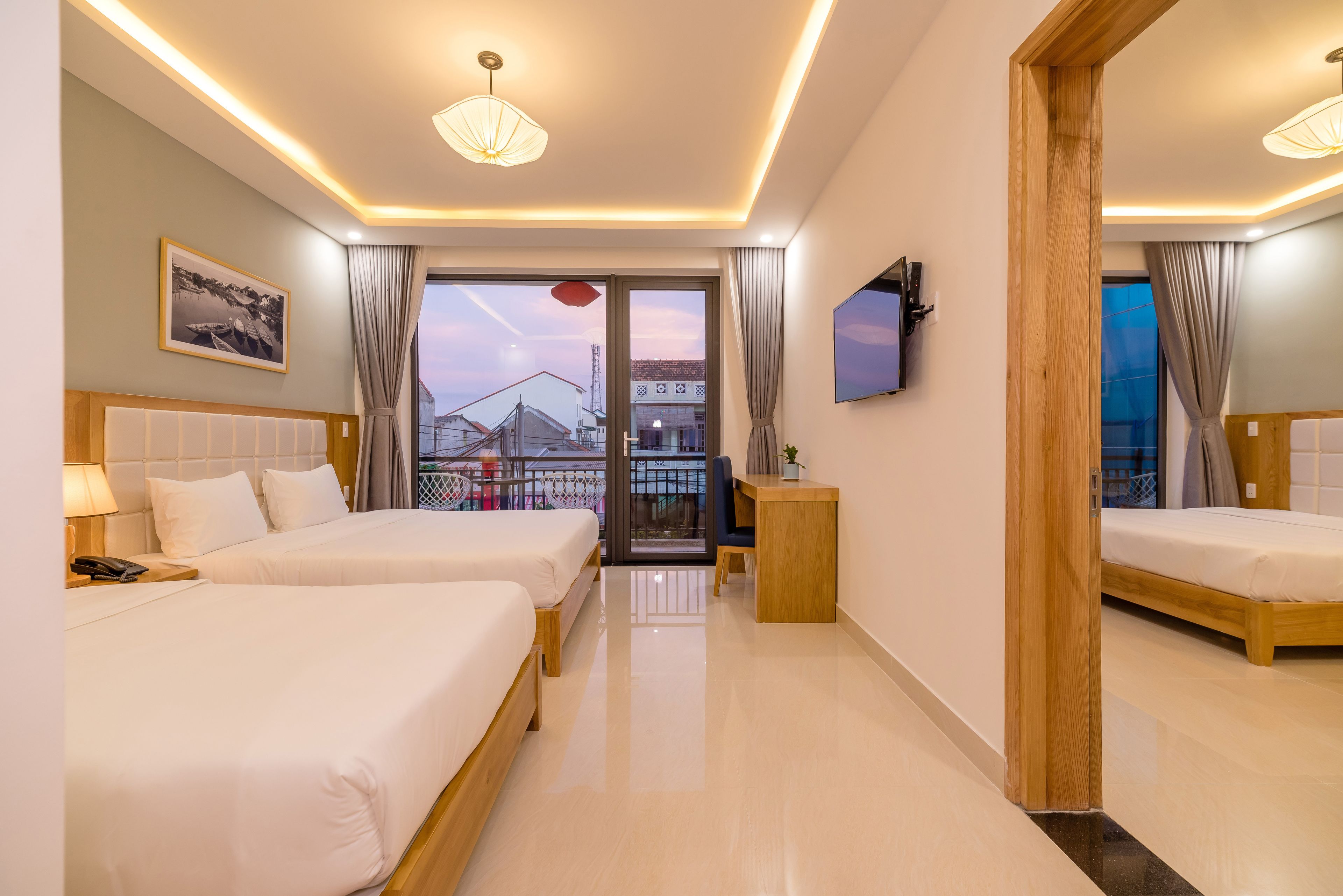 A hotel room with two beds, a desk, a TV, and a balcony with a view of buildings.