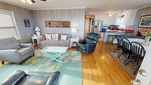 A living room with a grey sofa, a black leather chair, and a small table. There is a kitchen area with white cabinets and a red countertop. - Ocean Front at the Washout~ Foose's Folly (Folly Beach)