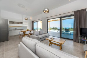Una sala de estar moderna con sofá, mesa de centro y comedor. Se observa una cocina con armarios blancos y un amplio ventanal con vistas a un balcón.