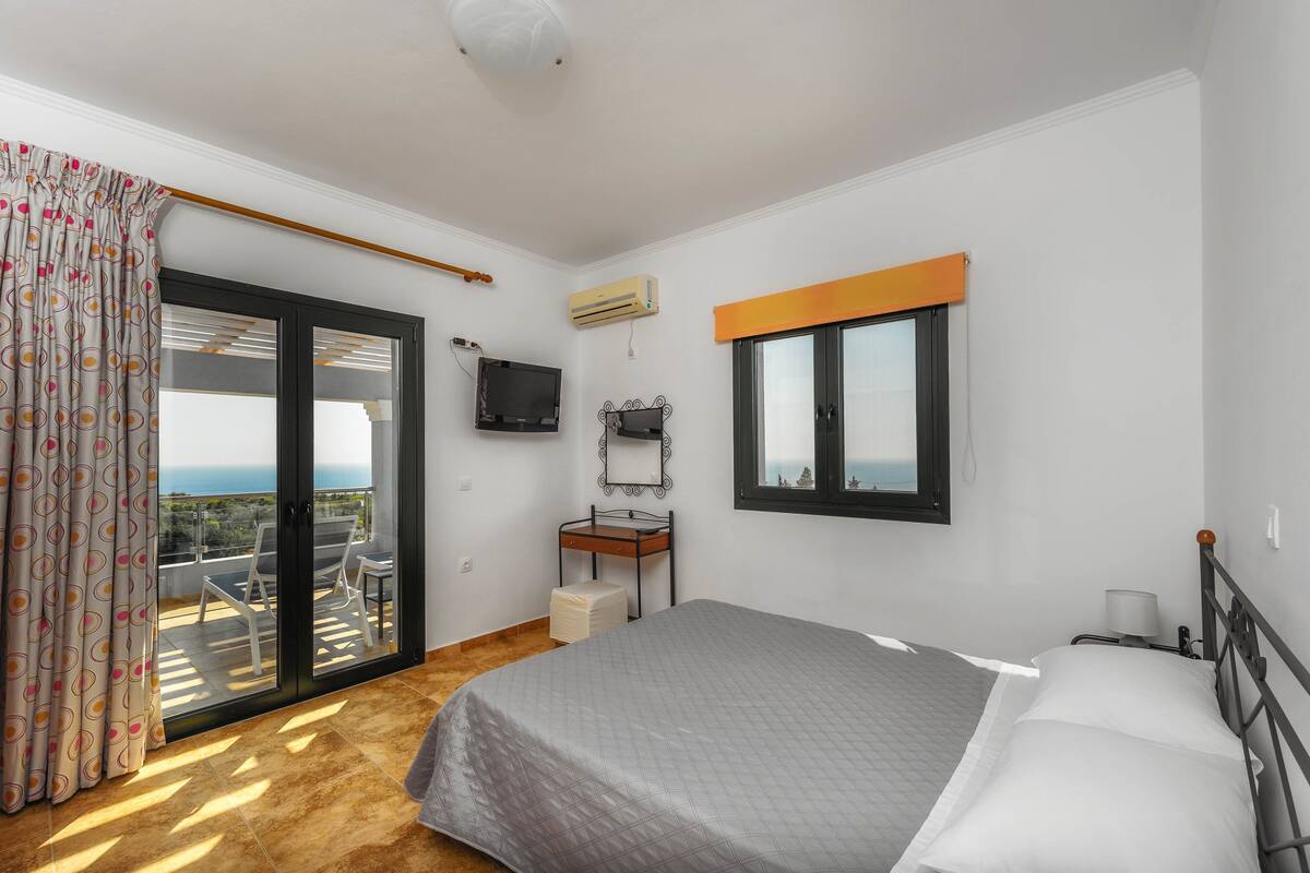 A bedroom with a bed, a small bedside table, a window, a TV, and a balcony with a view.