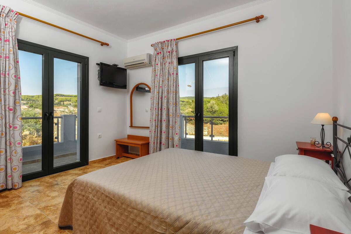 A bedroom with a bed, a TV, a small table, a lamp, and a view of the outdoors through a balcony door.