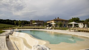 Une piscine aux bords ondulés, entourée de chaises longues et de parasols, avec des maisons en arrière-plan.