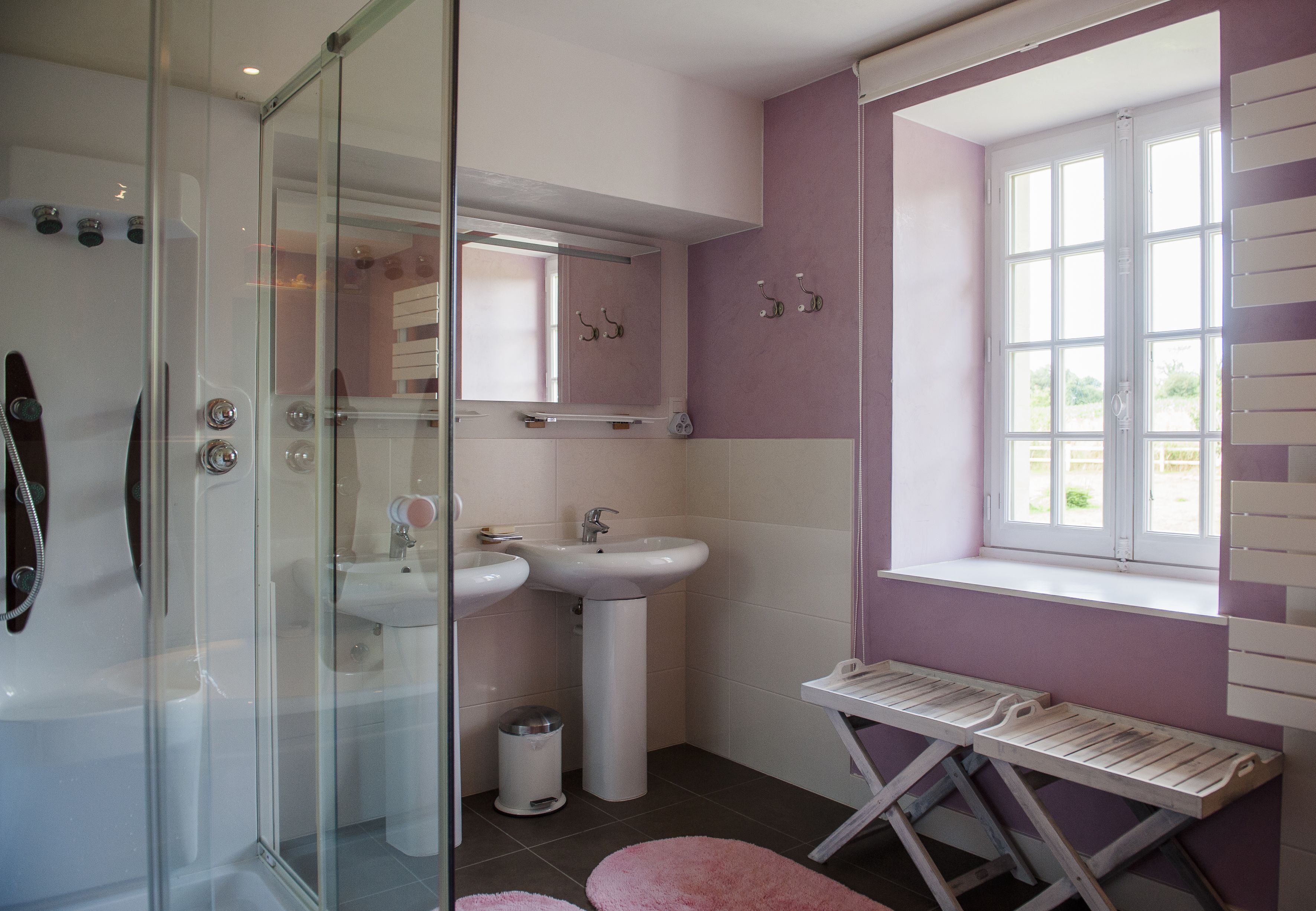 A bathroom with a glass shower enclosure, two sinks, a vanity with a mirror, and a window with blinds.